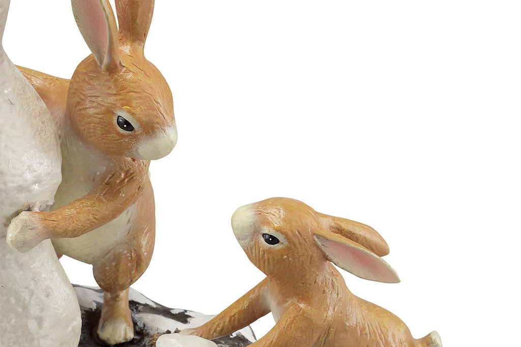 A close up of a sculpture of a pair of two bunnies building a bunny shaped snow man, while a cardinal bird hovers above.