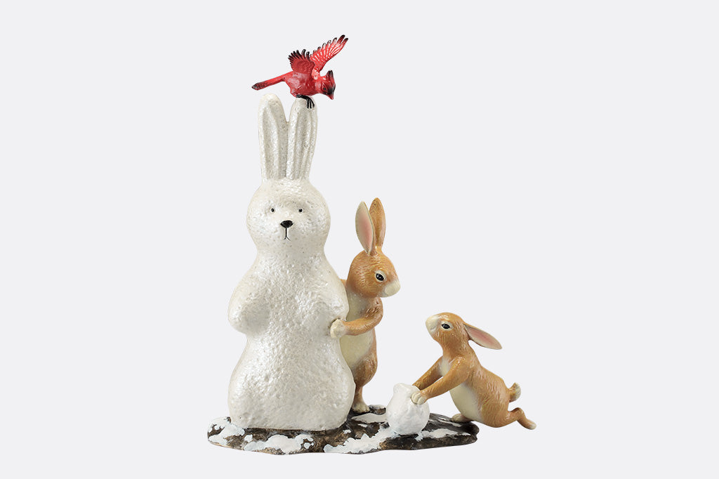 A sculpture of a pair of two bunnies building a bunny shaped snow man, while a cardinal bird hovers above.