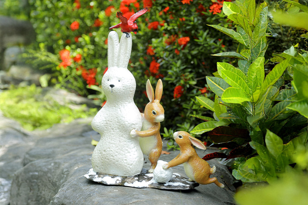 Two rabbits and a snowbunny statues on a rock with greenery and flowers in the background