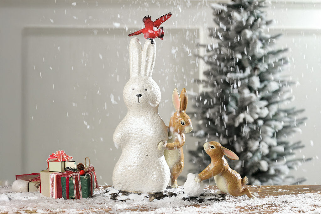 Decorative scene with a white rabbit figurine, small brown rabbits, and gift boxes in front of a Christmas tree.