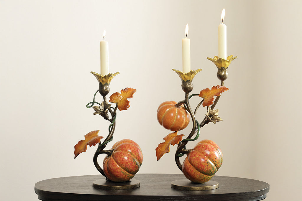 A candleholder statue of a vine blooming two pumpkins placed next to another candle holder statue of a vine blooming with only 1 pumpkin.