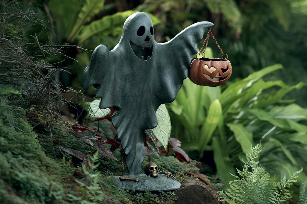 cast metal smiling ghost holding jack o lantern with candle inside set in outdoor garden space  