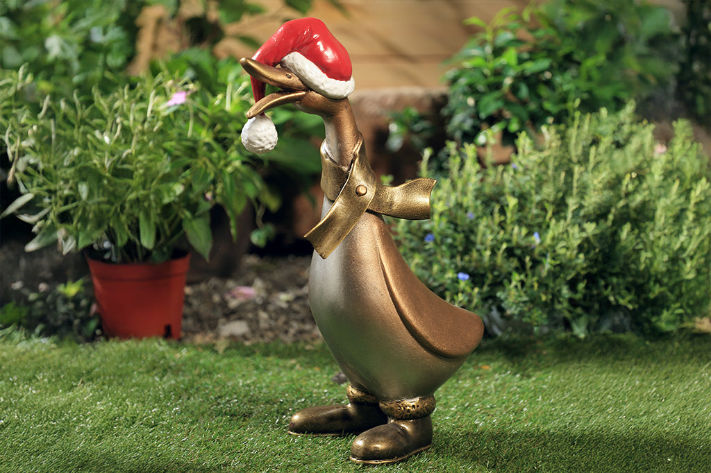 The side of a statue of a duck wearing a scarf, boots, and oversized santa hat covering his eyes placed in a garden.