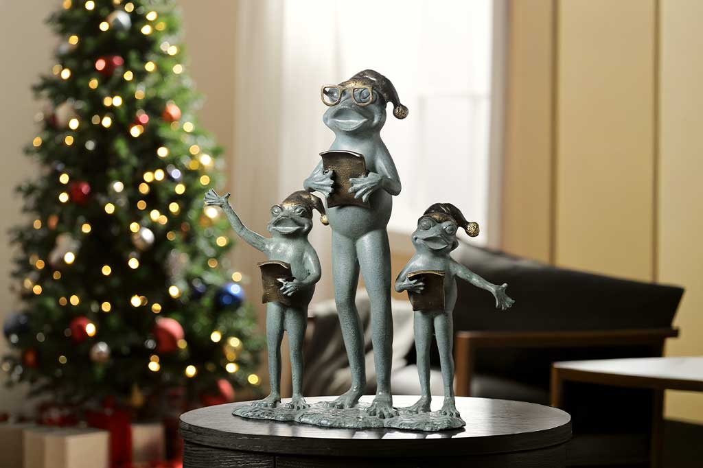 garden statue of 3 frogs with santa hats; they hold music as they carol in front of a decorated Christmas tree