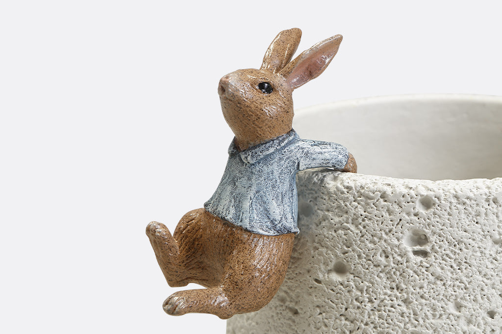 A hanging sculpture of two bunnies wearing blue shirts that dangle off of plant pots.