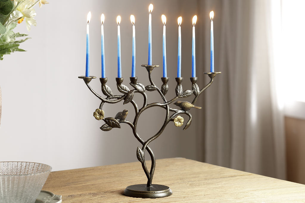 menorah with lit candles on a wooden surface. Features a cast metal arrangement of birds and blossoms on a branch and vine.
