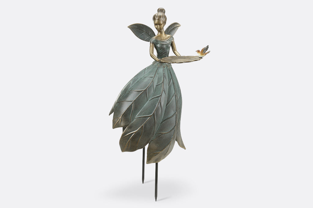 Cast metal garden statue of a fairy with leaf-like wings and dress, held up by removable stakes. shown on white background. Holds a bird feeder shaped like blossom and adorned with bird