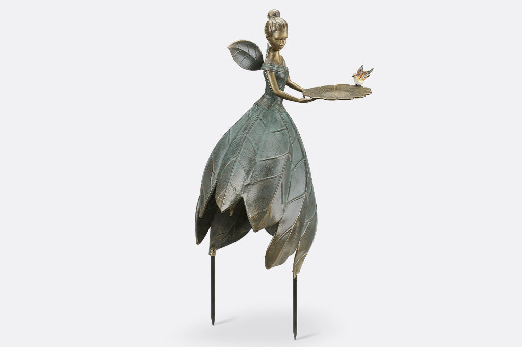 Cast metal garden statue of a fairy with leaf-like wings and dress, held up by removable stakes. shown on white background. Holds a bird feeder shaped like blossom and adorned with bird