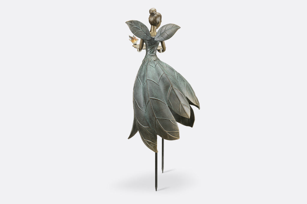 Cast metal garden statue of a fairy with leaf-like wings and dress, held up by removable stakes. shown on white background. Holds a bird feeder shaped like blossom and adorned with bird