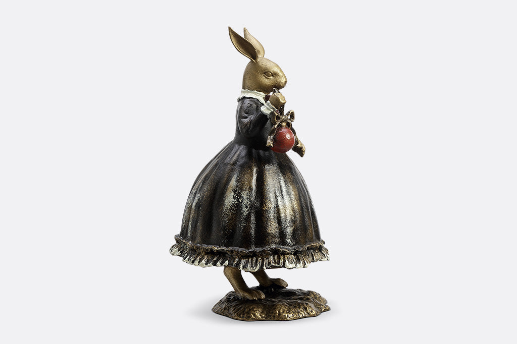 Sculpture of a bunny dressed in Victorian Era attire holding a red Chirstmas ornament. 
