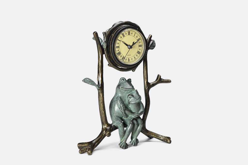 A clock with a sculpture of two frogs hugging while sitting on branches.