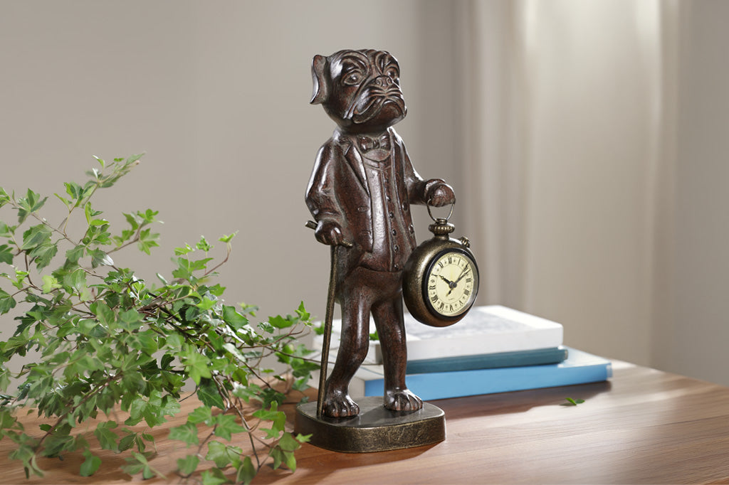 Bronze statue of a dog with a pocket watch on a wooden surface with a plant and books in the background.