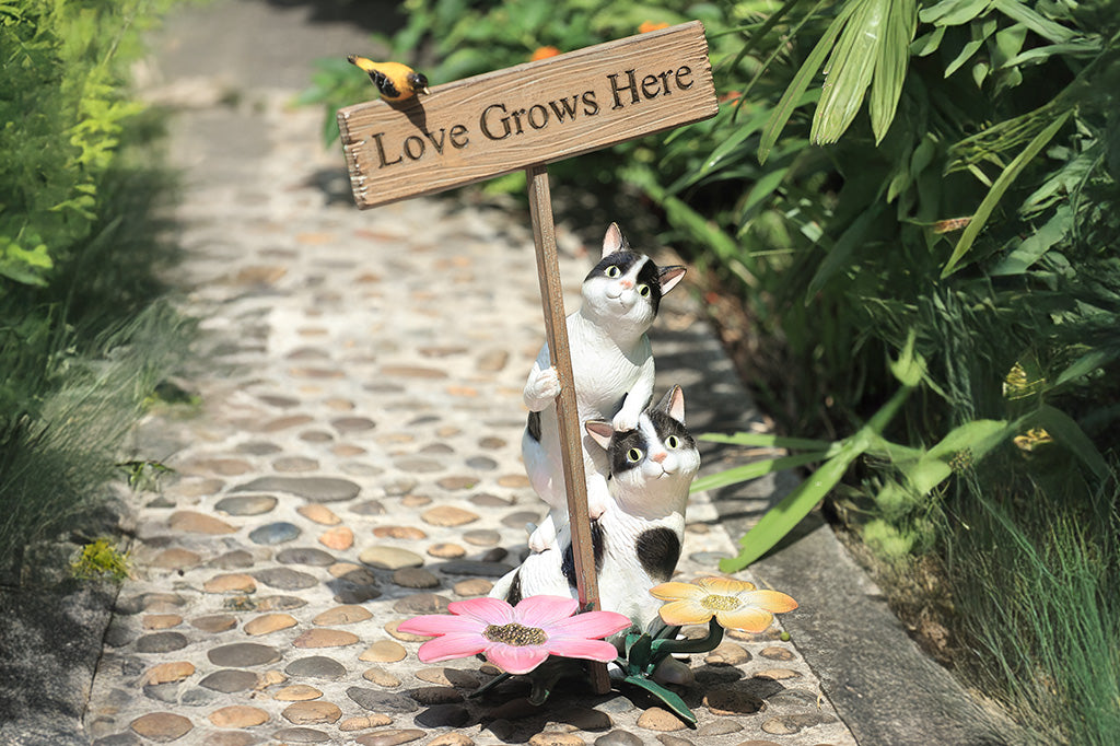 A garden sculpture of two cats holding a sign that says "love grows here" with a bird sitting on the sign and a pair of flowers at the bottom.