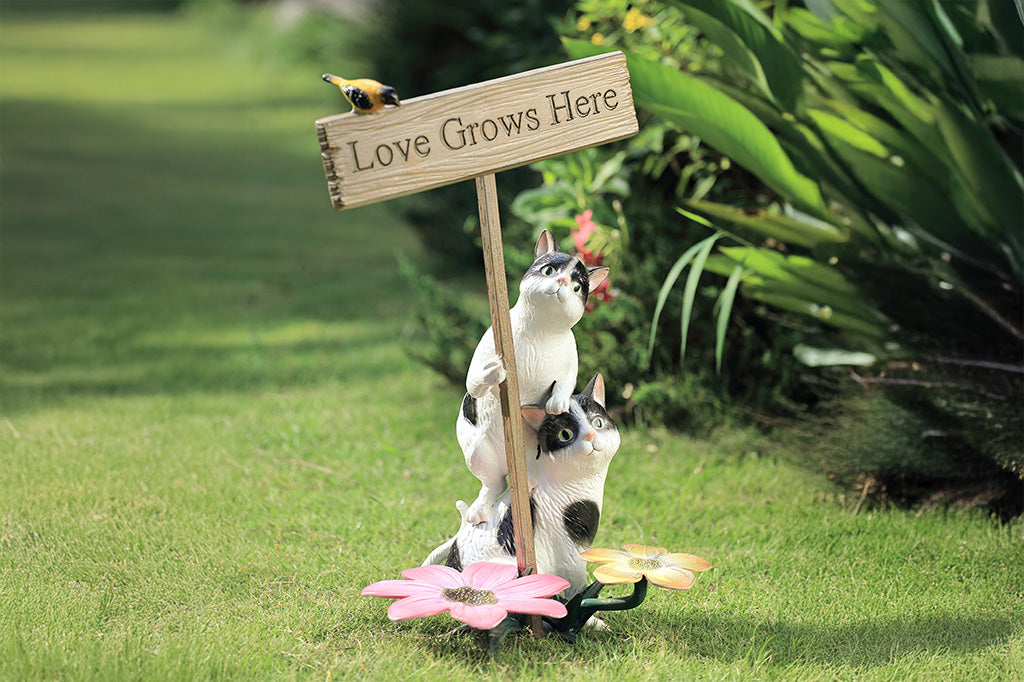 A garden sculpture of two cats holding a sign that says "love grows here" with a bird sitting on the sign and a pair of flowers at the bottom.