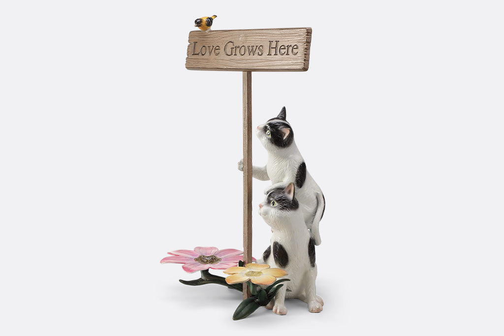 A garden sculpture of two cats holding a sign that says "love grows here" with a bird sitting on the sign and a pair of flowers at the bottom.