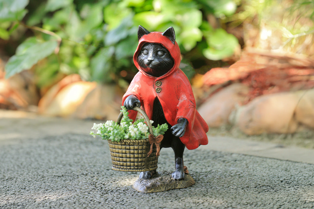 Cat figurine in a red hood holding a basket of flowers on a stone surface with greenery in the background.