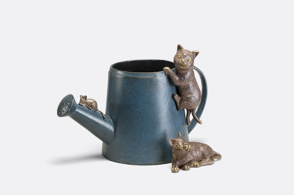 A decorative watering can planter with two cats climbing around to follow a mouse.