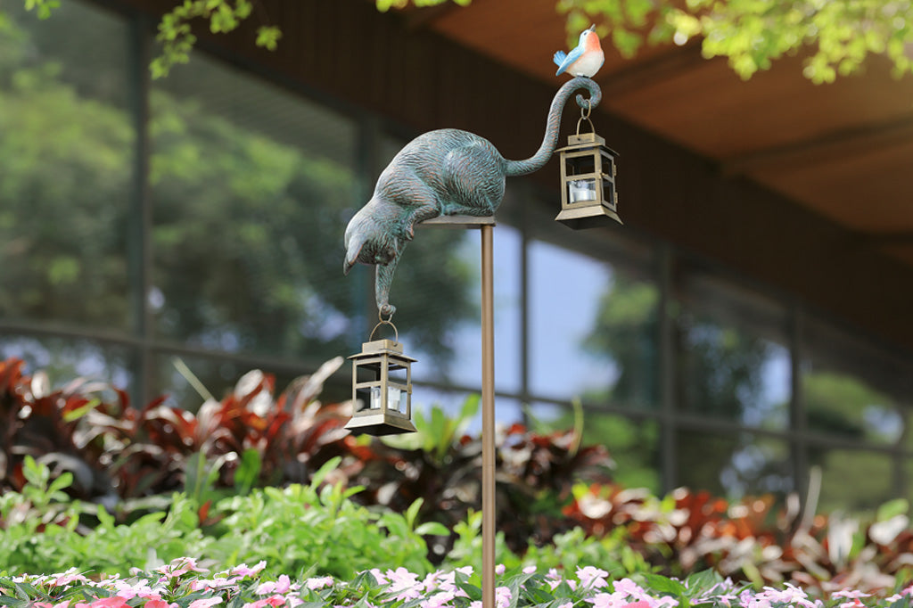 Decorative garden sculpture of a cat with a bird on its back, standing on a pedestal with lanterns, in a garden setting.
