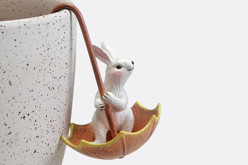 Pot hanger sculptures of two white bunnies in amber colored umbrellas. One bunny is facing left and the other is facing straight ahead.