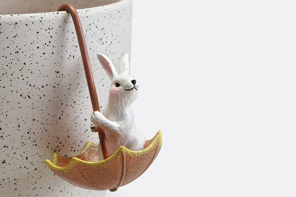 Pot hanger sculptures of two white bunnies in amber colored umbrellas. One bunny is facing left and the other is facing straight ahead.