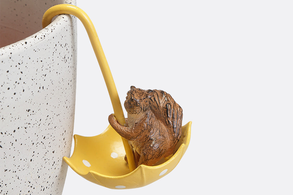 A hanging pot sculpture of a squirrel playing with a yellow umbrella.