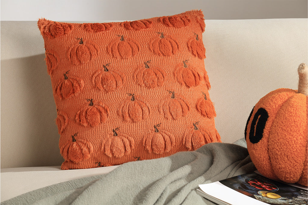 Orange pillow with pumpkin pattern on a bed next to a small pumpkin decoration.