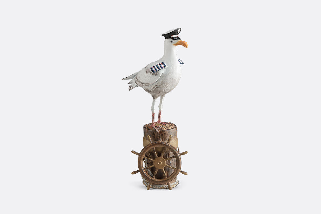 A sculpture of a seagull dressed in Captain regalia standing atop a dock adorned with rope and a ship's steering wheel.