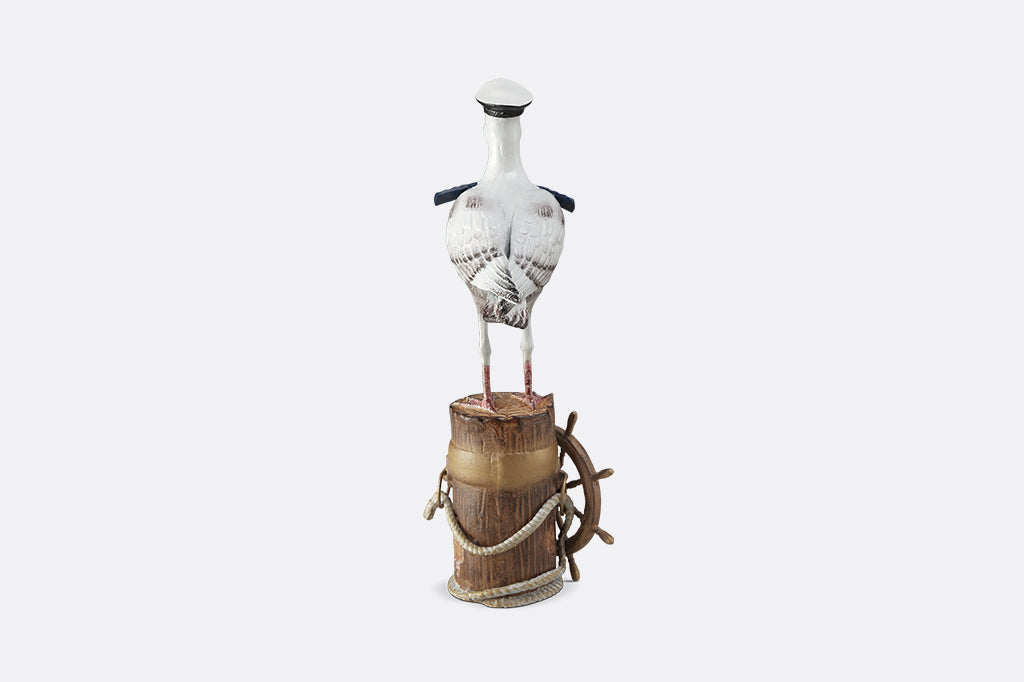 A sculpture of a seagull dressed in Captain regalia standing atop a dock adorned with rope and a ship's steering wheel.