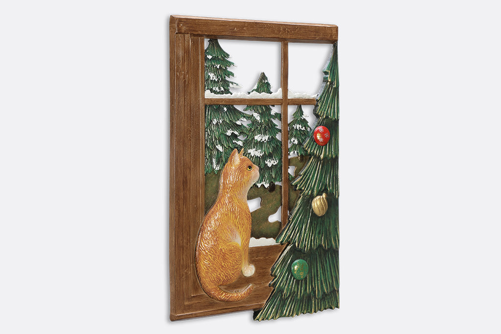 A decorative wall art of a cat looking out a window at trees next to a Chirstmas tree.