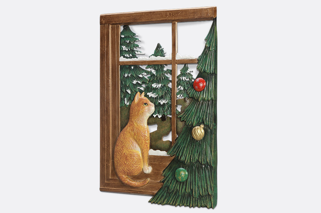 A decorative wall art of a cat looking out a window at trees next to a Chirstmas tree.