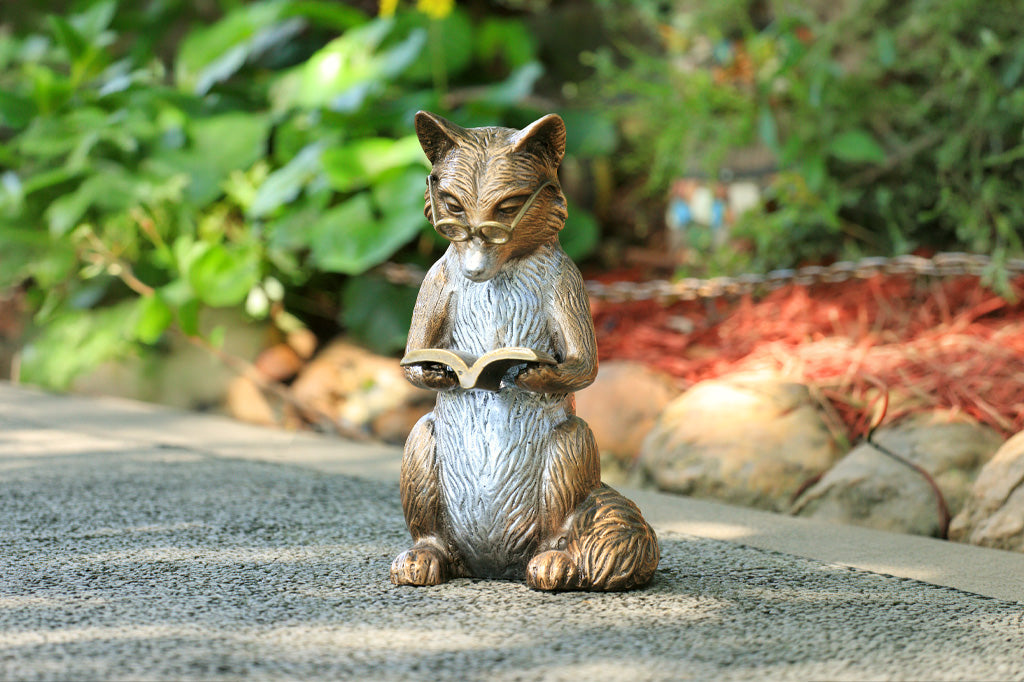 Statue of a fox reading a book on a stone path with greenery in the background