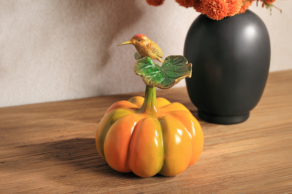 Decorative pumpkin with a bird on top on a wooden surface next to a black vase with flowers.