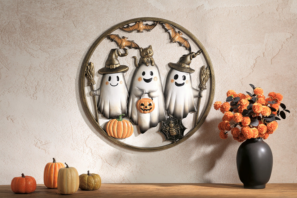 Decorative wall piece with ghosts, pumpkins, and bats on a neutral wall with pumpkins and a vase on a table.