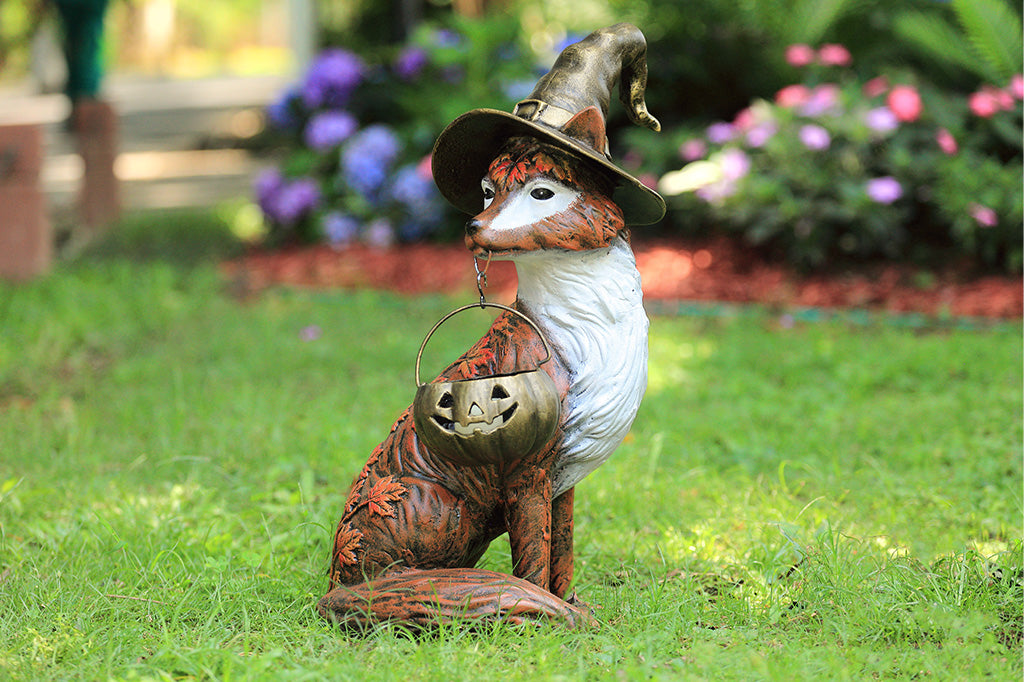 Garden statue of a fox wearing a witch hat with a pumpkin, sitting on grass with flowers in the background.