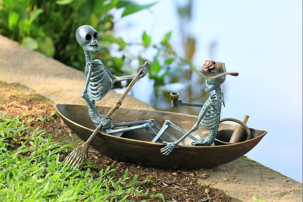Two skeleton figures in a boat with one rowing and the other playing a harmonica, set against a natural background.
