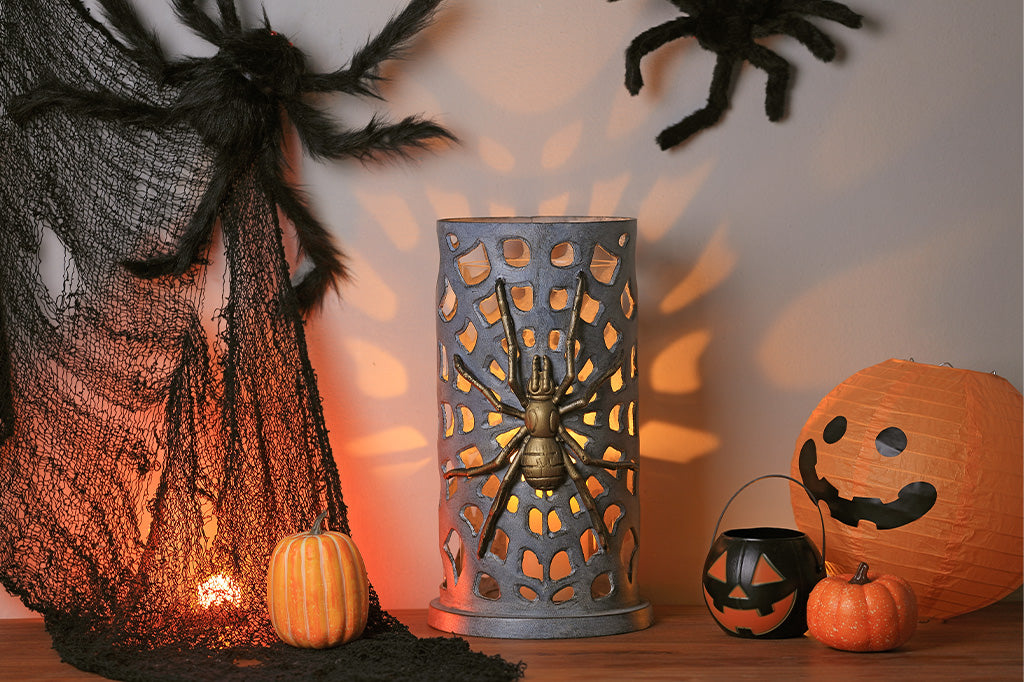 Halloween decoration display featuring a tall metal spider lantern in the center, a black web curtain and fake spider on the left, a jack-o'-lantern face on the right, small pumpkins, and a black cauldron with a bucket, arranged on a wooden surface against a wall with spiderweb shadows.