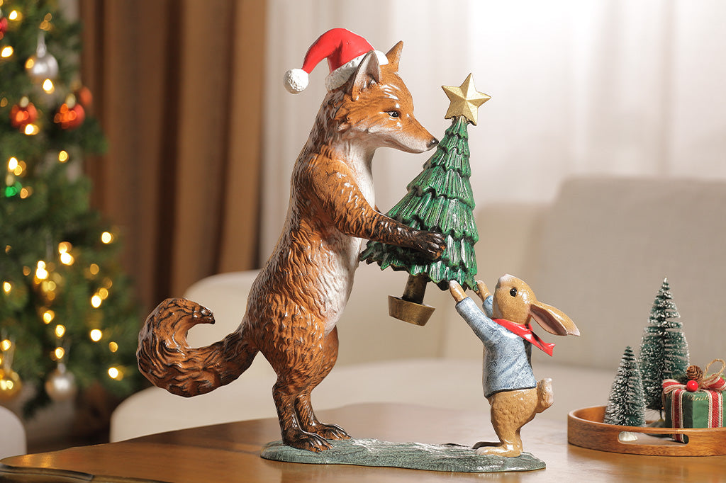 A sculpture of a fox wearing a santa hat is giving a rabbit with a scarf a christmas tree with a star on top.