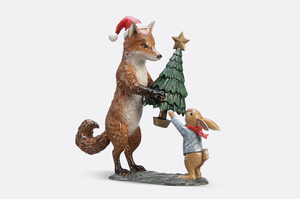 A sculpture of a fox wearing a santa hat is giving a rabbit with a scarf a christmas tree with a star on top.