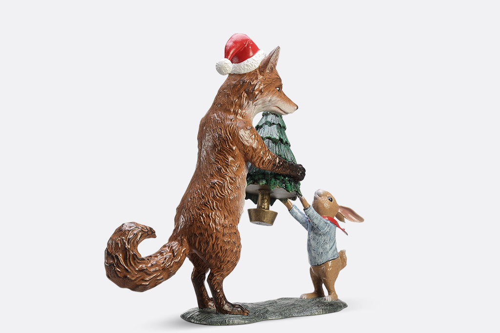 A sculpture of a fox wearing a santa hat is giving a rabbit with a scarf a christmas tree with a star on top.