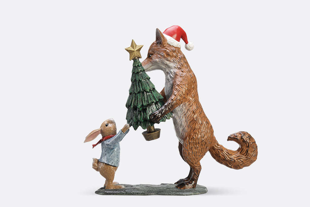 A sculpture of a fox wearing a santa hat is giving a rabbit with a scarf a christmas tree with a star on top.