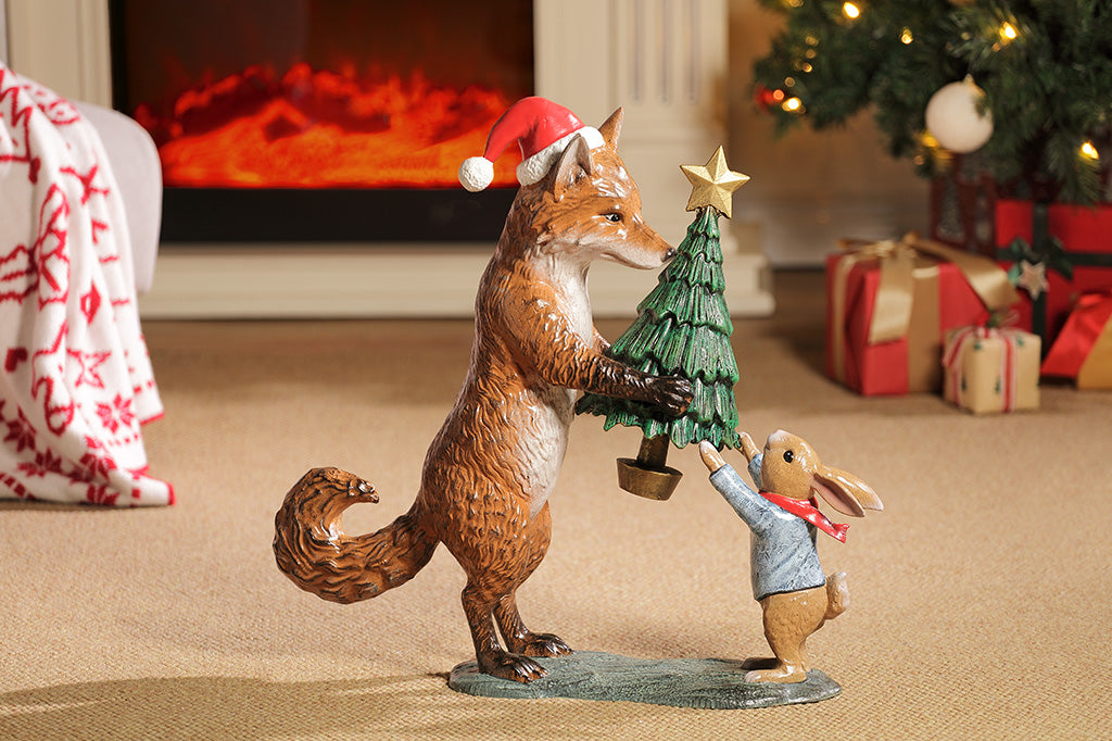 A sculpture of a fox wearing a santa hat is giving a rabbit with a scarf a christmas tree with a star on top.