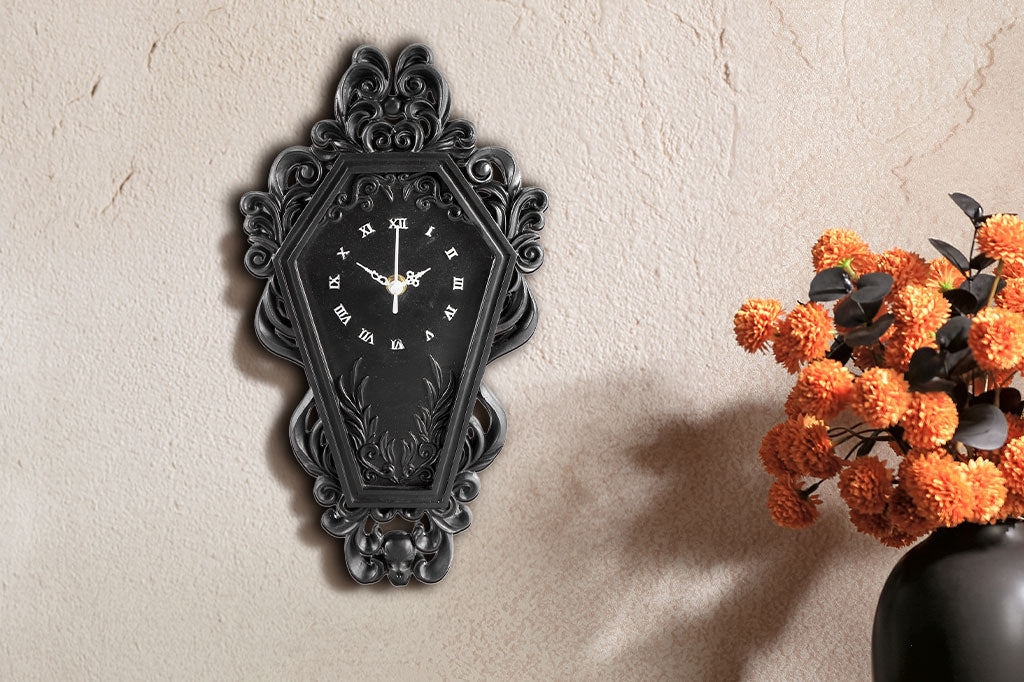Gothic coffin-inspired wall clock with intricate design on a light beige wall next to a vase with orange flowers.