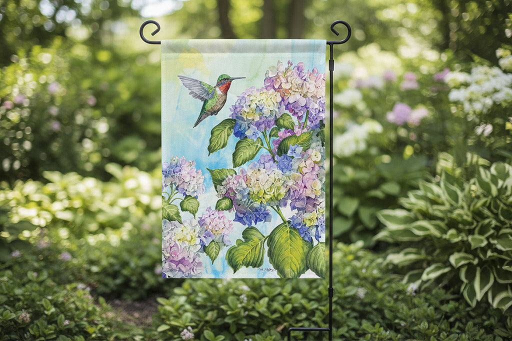 house decor flag with a hummingbird and hydrangea design shown on iron staked flag pole in a garden