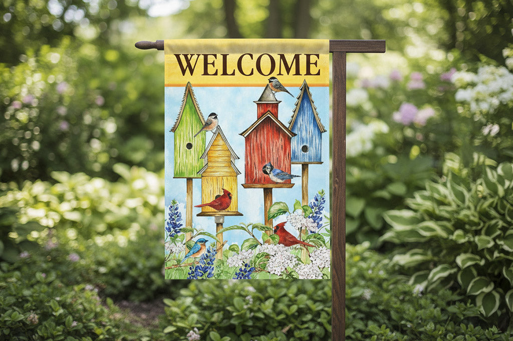garden decor flag that says "Welcome" on yellow; a variety of songbirds and birdhouses amongst blooming bluebonnets shown as the main design. Shown on wood flag holder in a garden