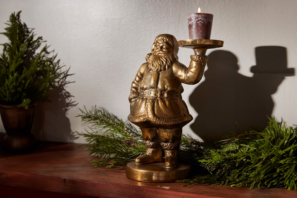 Metal sculpture of Santa holding a votive candle is  upon a wood fireplace mantle with evergreens around