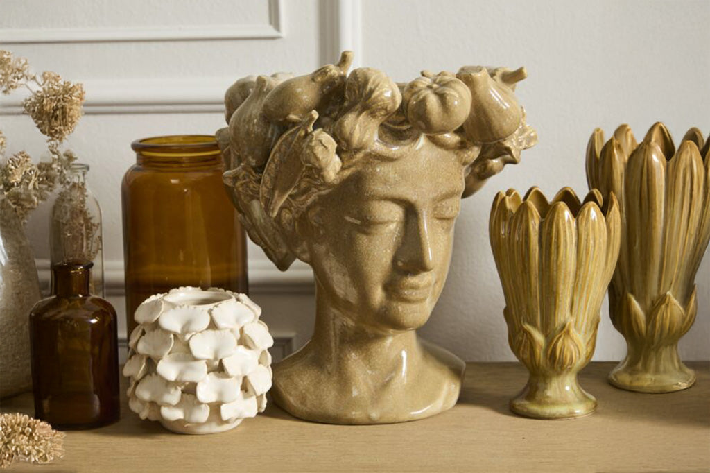 Decorative ceramic pieces including a ceramic vase shaped as a woman's bus with cornucopia wreath and various vases on a wooden surface.