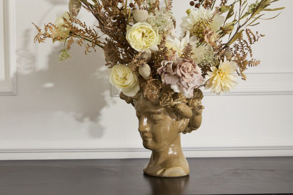 Decorative vase shaped like a head with cornucopia wreath holds a floral arrangement against a white wall.