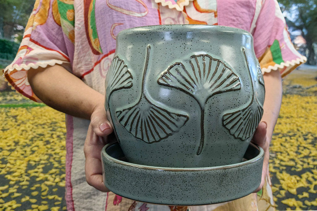 Person holding a gray-green speckled stoneware planter with leaf designs outdoors.