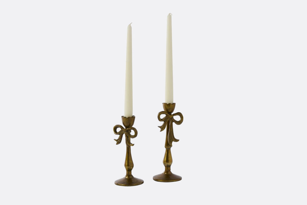 A pair of ornate brass candlesticks with white taper candles, set against a plain light gray background.