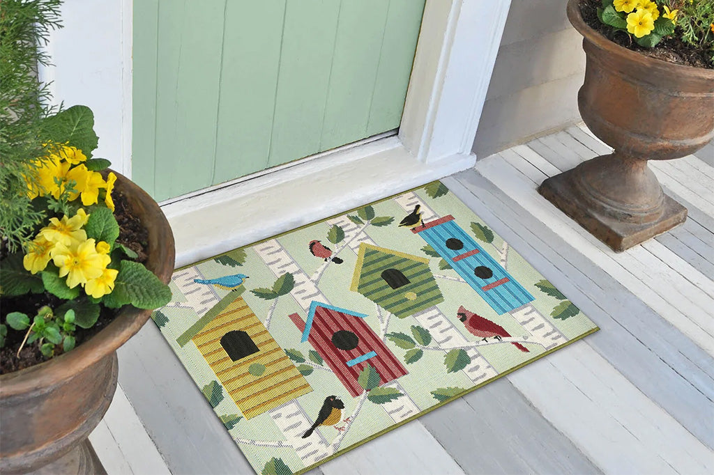 Decorative doormat with birdhouses and birds on a wooden floor, flanked by potted plants.